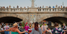 Grafika przedstawia ludzi stojących w okręgu i trzymających się za ręce. W tle most. Słoneczna pogoda, lato. 