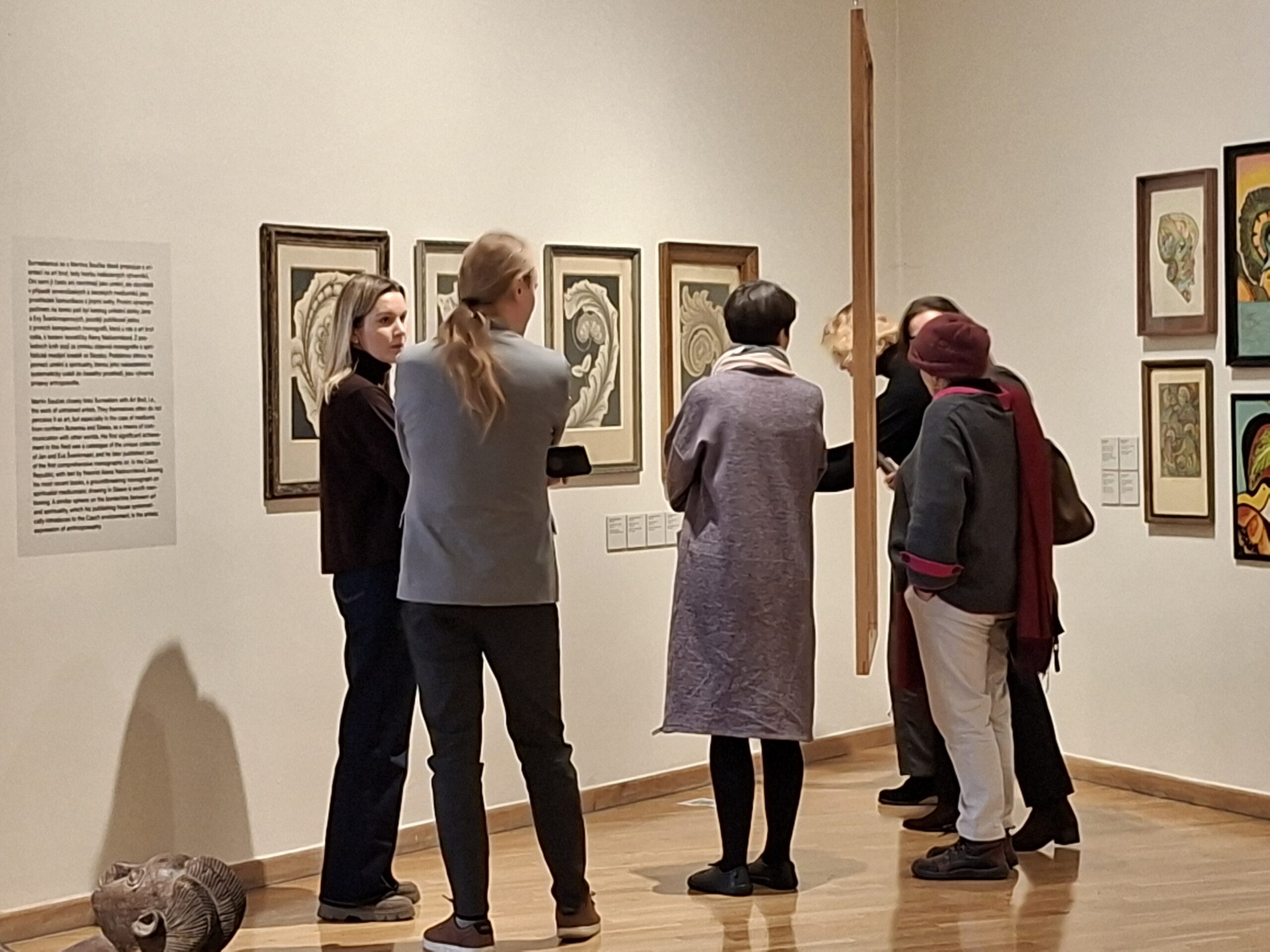 Delegacja MCK ogląda wystawę w Muzeum Sztuki w Ołomuńcu, kilka osób na tle grafik w róznych kolorach, wiszących w drewnianych ramach na białych ścianach.