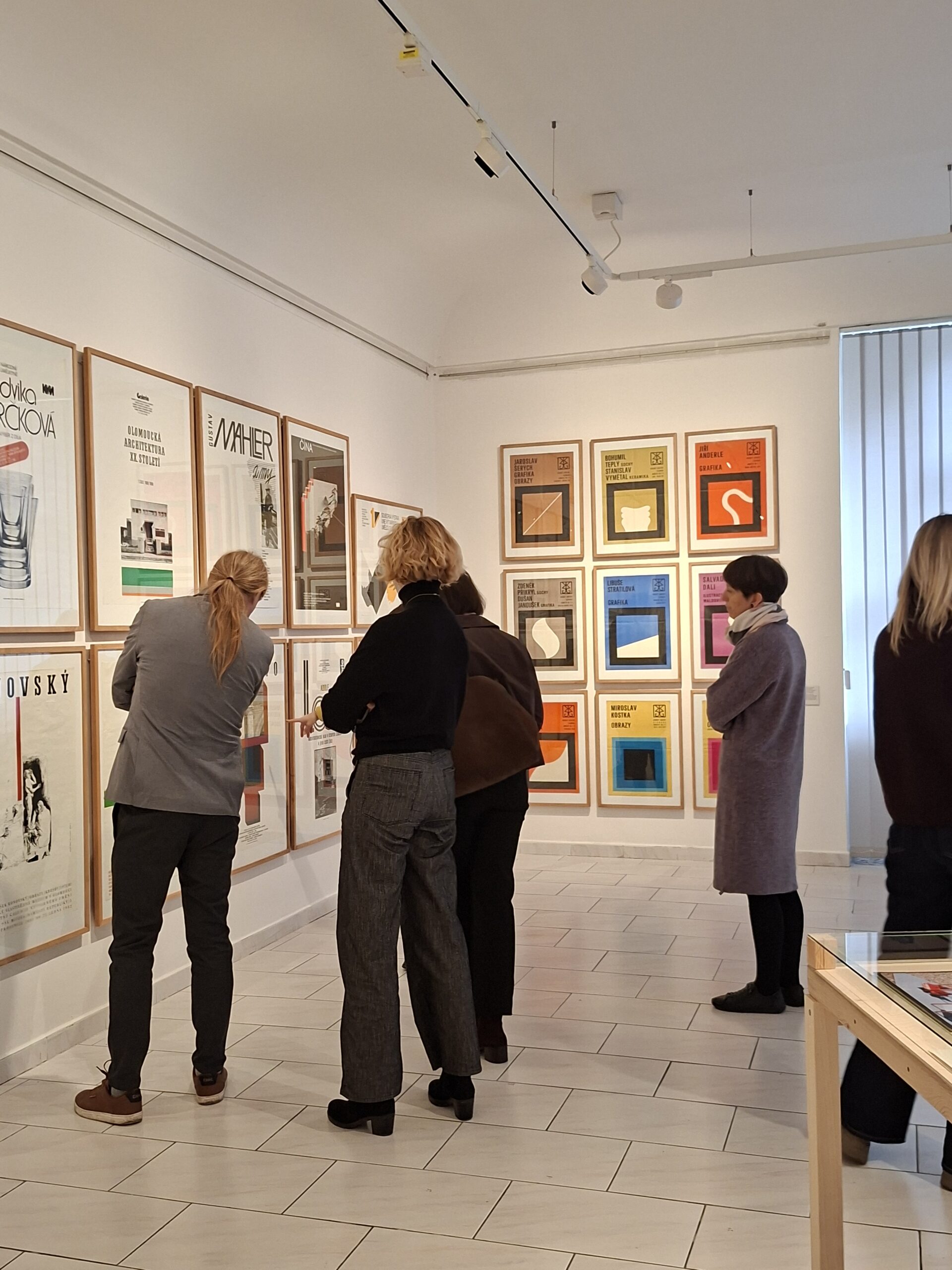 Delegacja MCK ogląda wystawę w Muzeum Sztuki w Ołomuńcu, kilka osób na tle grafik w róznych kolorach, wiszących w drewnianych ramach na białych ścianach.