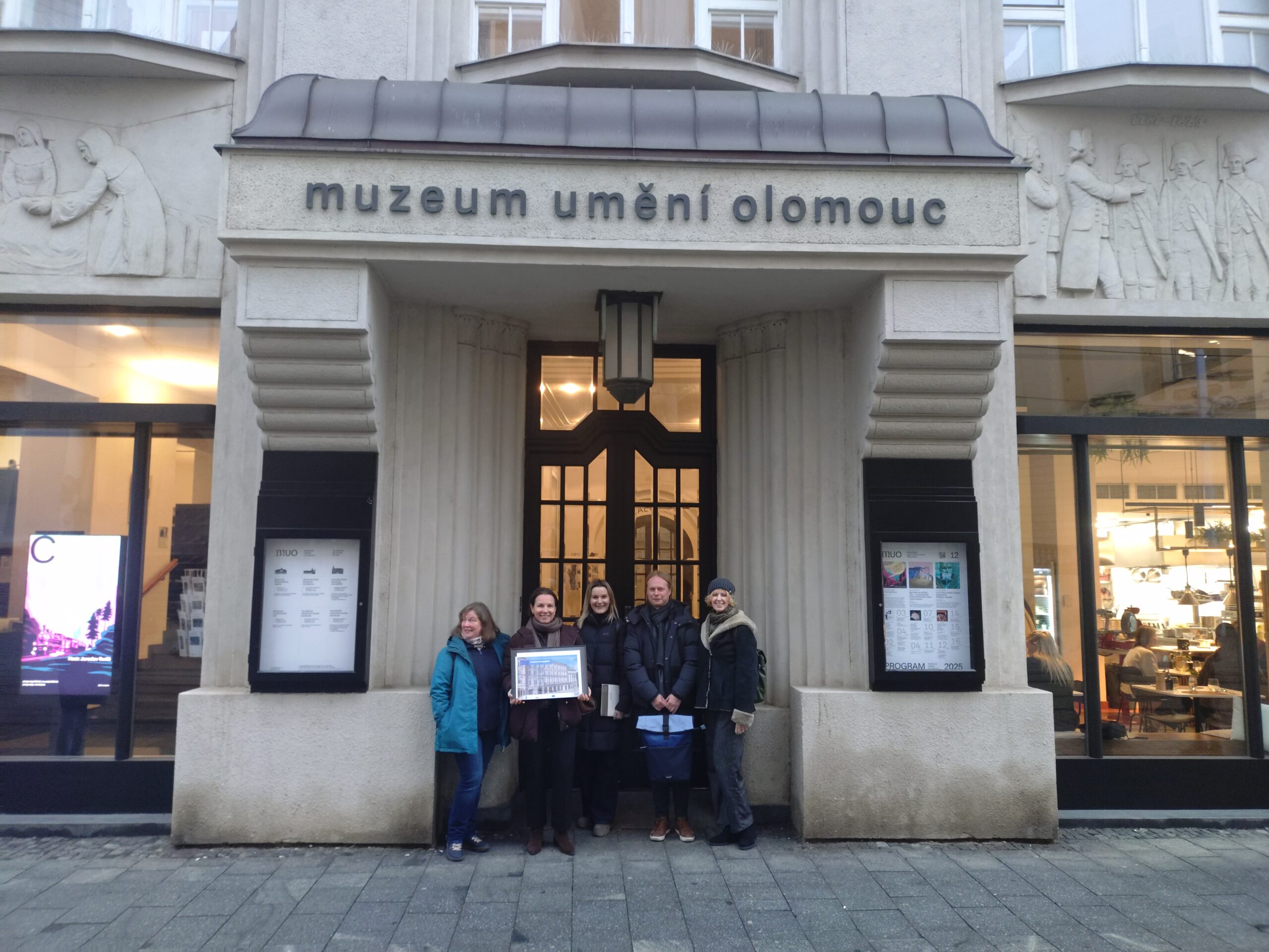 Osoby reprezentujące MCK na tle fasady Muzeum Sztuki w Ołomuńcu. Za nimi drewniane drzwi z przeszkleniem, po boku tablice informacyjne, osoba z lewej strony trzyma tablicę ze zdjęciem siedziby MCK.