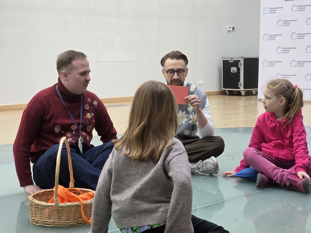 Dwoje prowadzących i dzieci siedzą w kręgu podczas warsztatów edukacyjnych z kolorami.