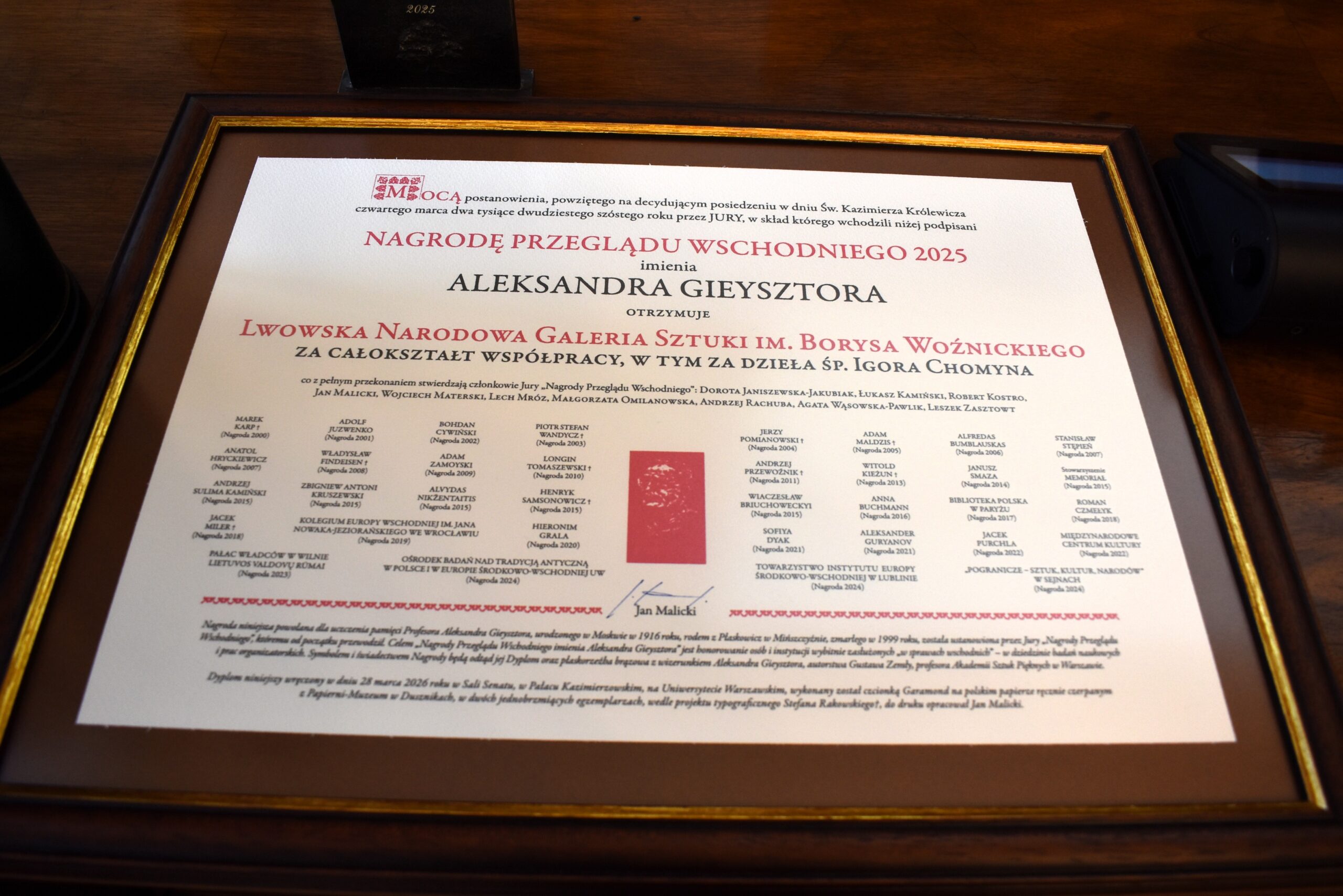 Oprawiony dyplom Nagrody Przeglądu Wschodniego 2025 im. Aleksandra Gieysztora dla Lwowskiej Narodowej Galerii Sztuki im. Borysa Woźnickiego, sfotografowany na drewnianym stole.