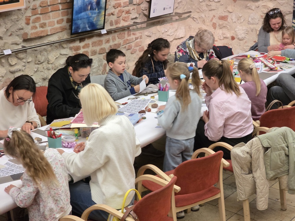 Zdjęcie z warsztatów rodzinnych w przestrzeni wystawowej. Dzieci i dorośli siedzą przy długim stole, wykonując prace plastyczne. W tle widoczne są ceglane i kamienne ściany oraz prezentowane obrazy.