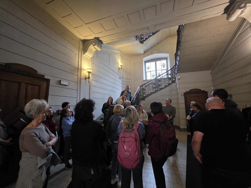 Zdjęcie wnętrza Domu pod Globusem w Krakowie. Uczestnicy wydarzenia stoją w holu przy reprezentacyjnych schodach z ozdobną balustradą i witrażowym oknem. W jasnym, zabytkowym wnętrzu widoczne są drewniane drzwi oraz dekoracyjne detale architektoniczne.