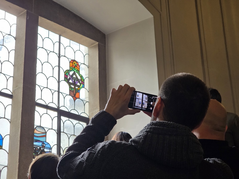 Photograph of the interior of the House Under the Globe in Krakow. Event participants are gathered near a large stained-glass window, while one person is photographing a stained-glass detail with a mobile phone. Historic architectural details are visible in the bright interior.