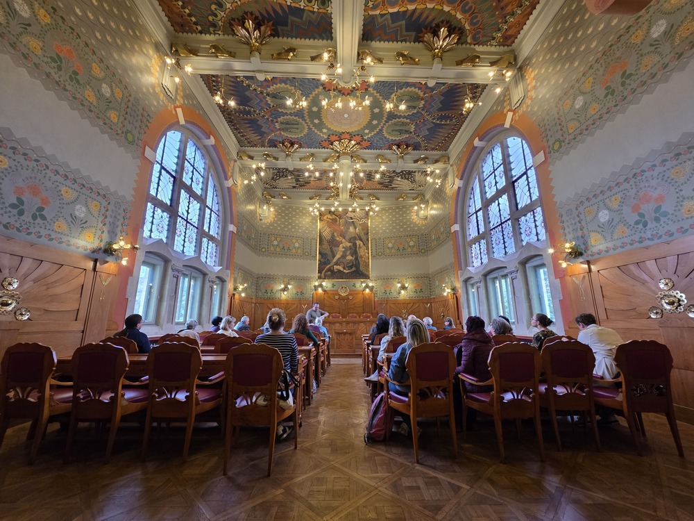 Photograph of the interior of the House Under the Globe in Krakow. In the richly decorated hall with a painted ceiling, ornamental walls, and large windows, participants are seated during a lecture or meeting. Dr Michał Wiśniewski is speaking at the front of the room.