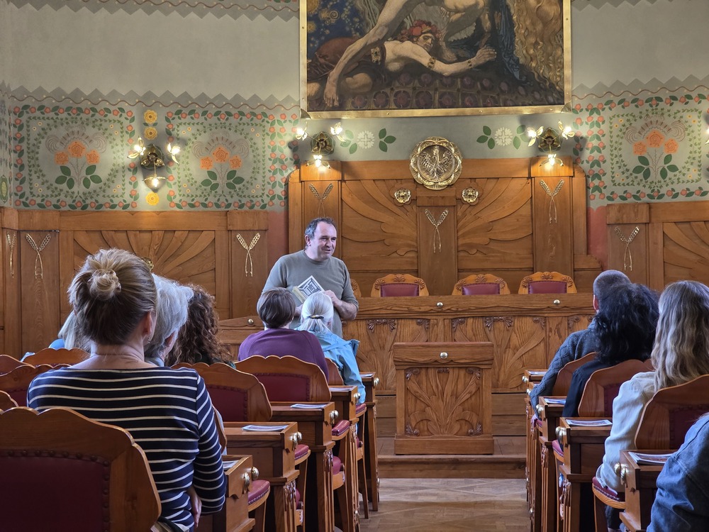 Photograph from a meeting at the House Under the Globe in Krakow. Dr Michał Wiśniewski is speaking to participants seated on wooden benches in a richly decorated hall with ornamental walls and Art Nouveau paintings.