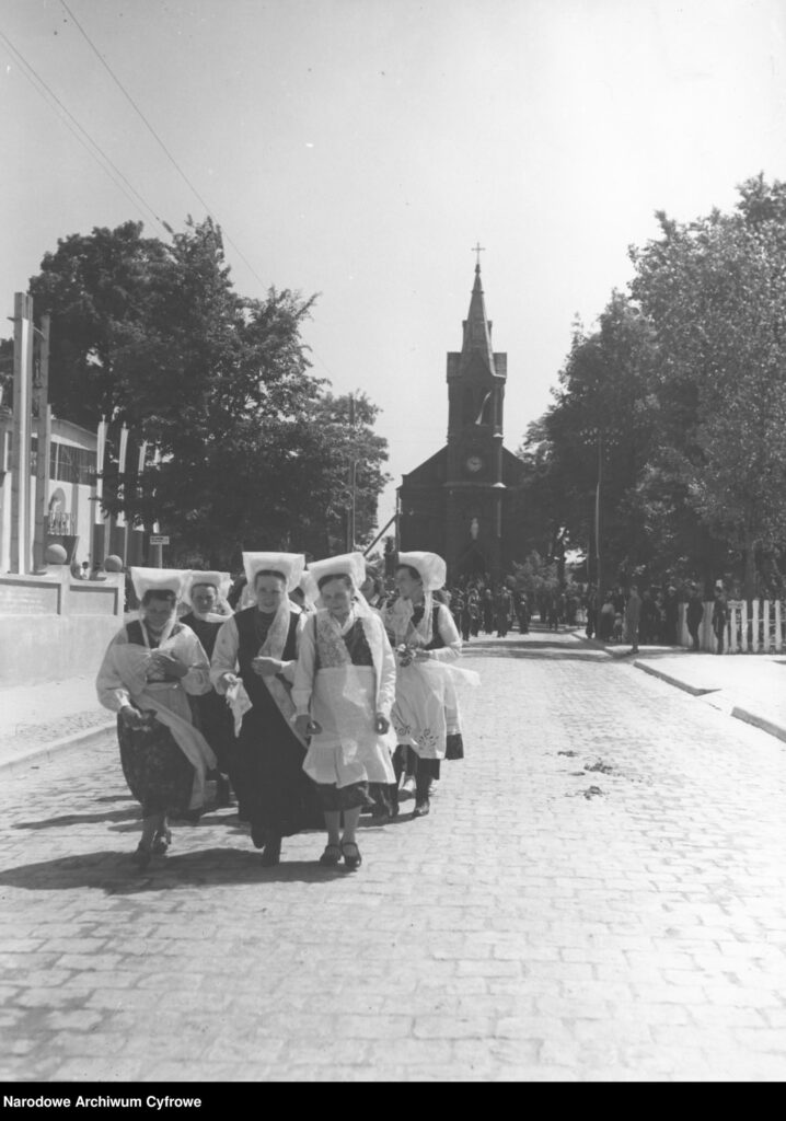 Kobiety idące drogą w strojach ludowych, za nimi widać sylwetkę kościoła.