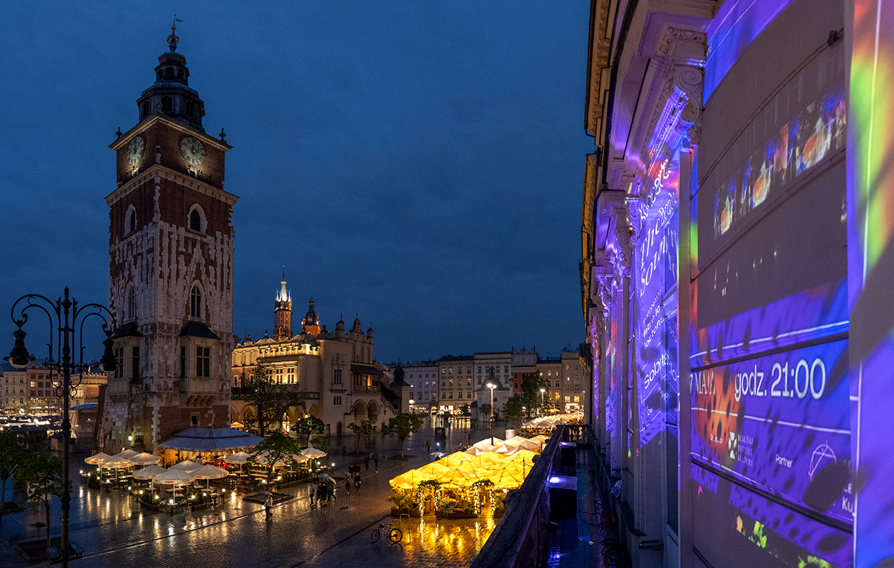 Rozświetlona fasada kamienicy i widok na Rynek Główny nocą. 