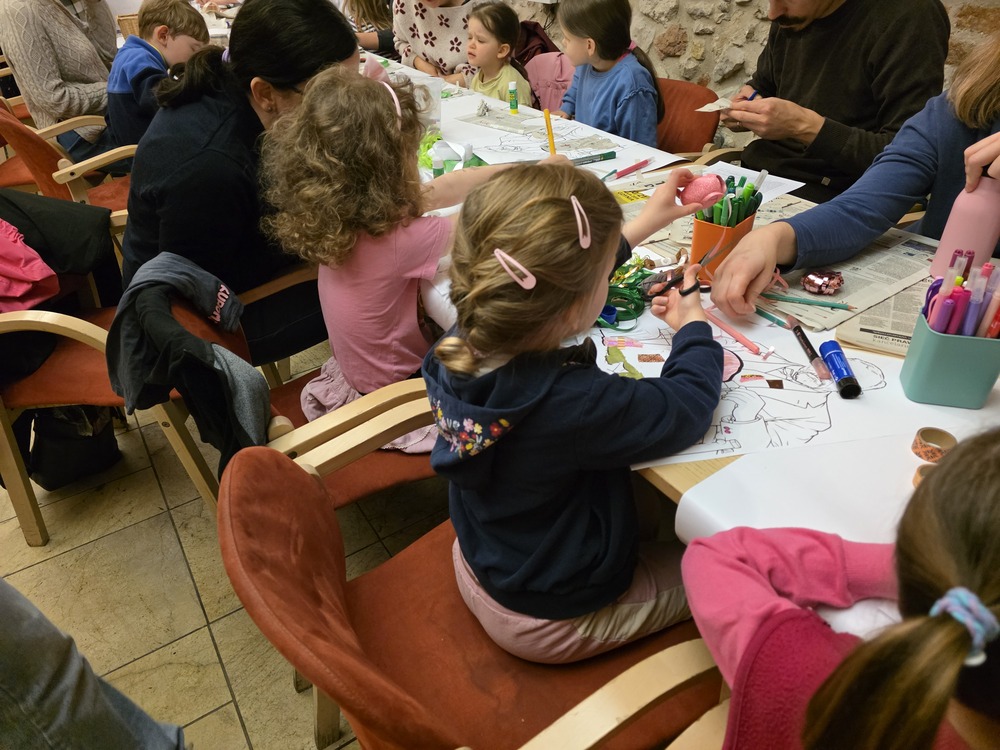 Photograph from a family art workshop for children. Participants are seated at a table creating artworks using paper, crayons, glue, and colourful materials. Stone walls of the interior are visible in the background.
