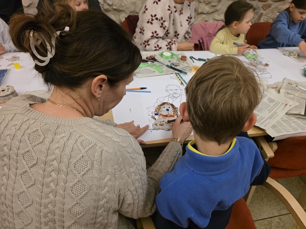 Photograph from a family workshop for children. An adult and a child are creating an artwork together at a table covered with paper, crayons, and newspaper cut-outs. Other workshop participants are visible in the background.