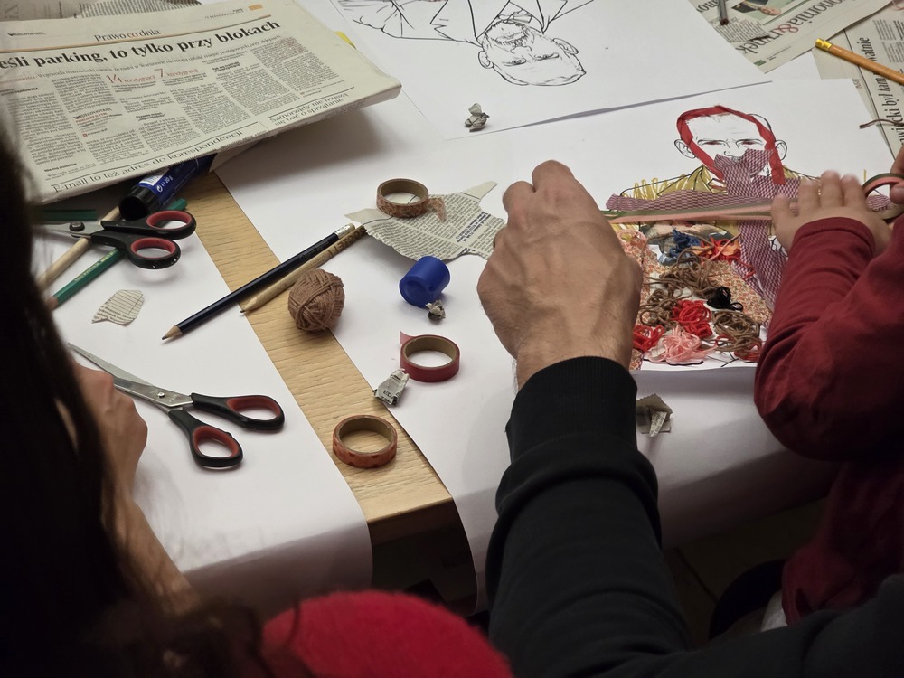 Close-up of a table during an art workshop. Children and adults are creating collages from paper, yarn, and newspaper cut-outs depicting portraits of figures. Scissors, crayons, and art materials are spread across the table.