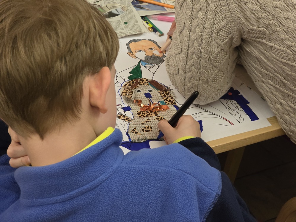 Close-up of a child taking part in an art workshop. Sitting at a table, the child creates a colourful artwork using paper, cut-outs, and drawings of figures. Art materials and the hand of an assisting adult are visible nearby.