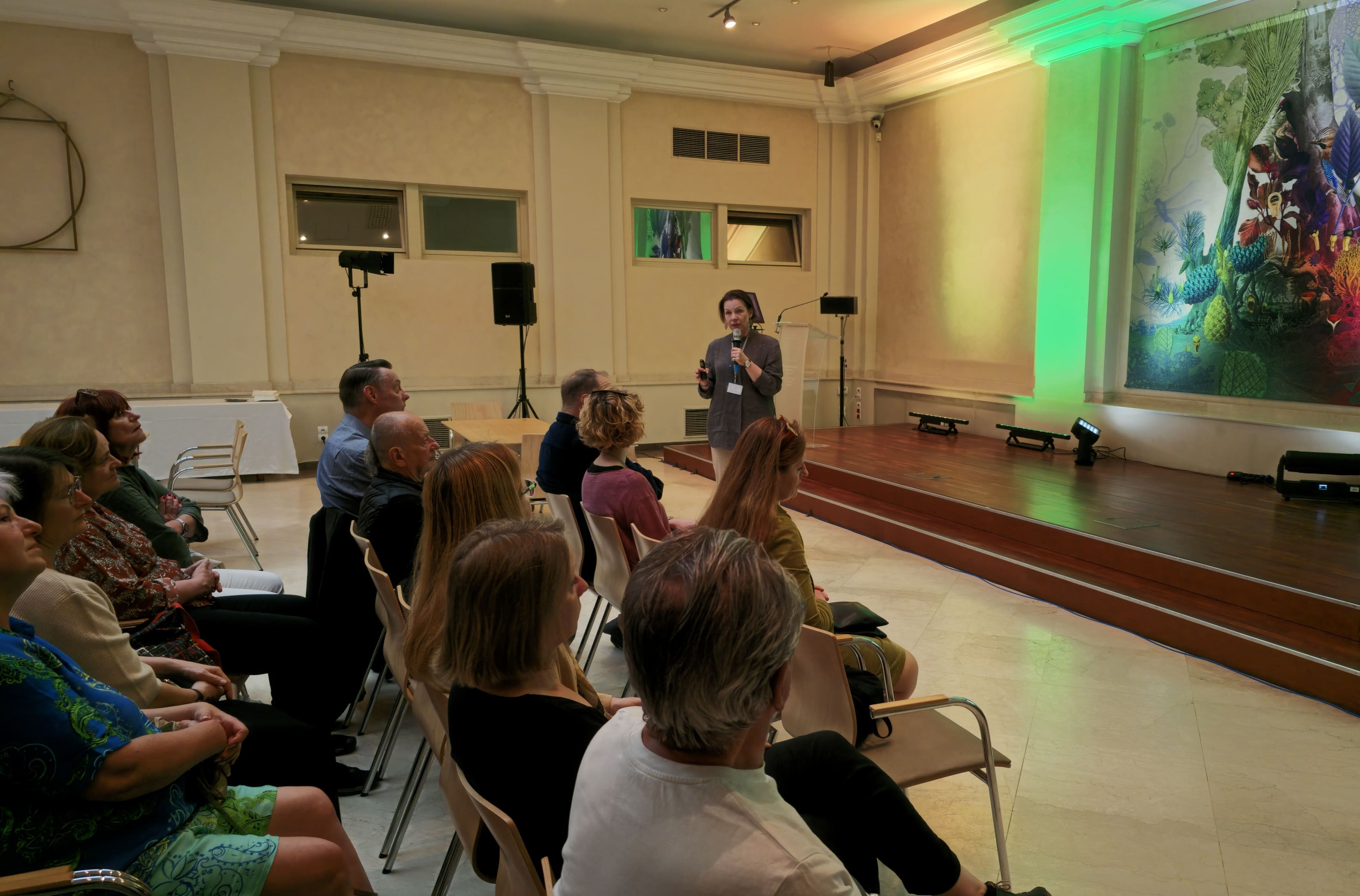 Spotkanie gości ICOM w sali konferencyjnej MCK. Uczestnicy siedzą przed sceną i słuchają prezentacji prowadzonej przez Agatę Wąsowską-Pawlik, która trzyma mikrofon. Wnętrze oświetlone jest zielonym światłem, a na ścianie widoczna jest wielkoformatowa, kolorowa praca artystyczna.