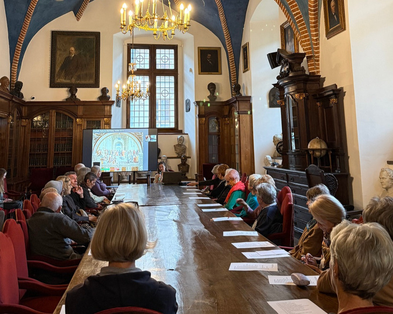 Photograph from the lecture A New View of the World: Print and Perspective. Participants are seated around a long table in a historic hall with wooden furniture, portraits, and chandeliers. A multimedia presentation displayed on a screen is visible during the event.