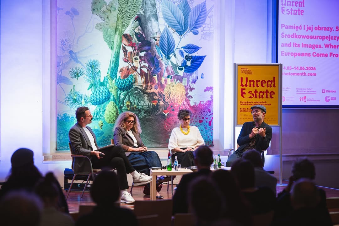 Debata podczas otwarcia 23. Miesiąca Fotografii w Krakowie w Międzynarodowym Centrum Kultury. Na scenie siedzą cztery osoby uczestniczące w rozmowie panelowej, a jedna z nich przemawia do publiczności. W tle widoczny jest plakat wydarzenia „Unreal Estate” oraz kolorowa dekoracja ścienna.