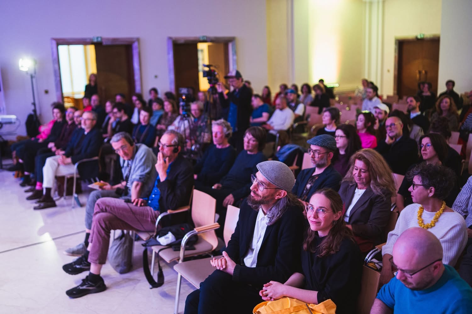 Publiczność zgromadzona podczas otwarcia 23. Miesiąca Fotografii w Krakowie w Międzynarodowym Centrum Kultury. Uczestnicy siedzą na widowni i słuchają wydarzenia, a w tle widoczna jest kamera rejestrująca spotkanie.