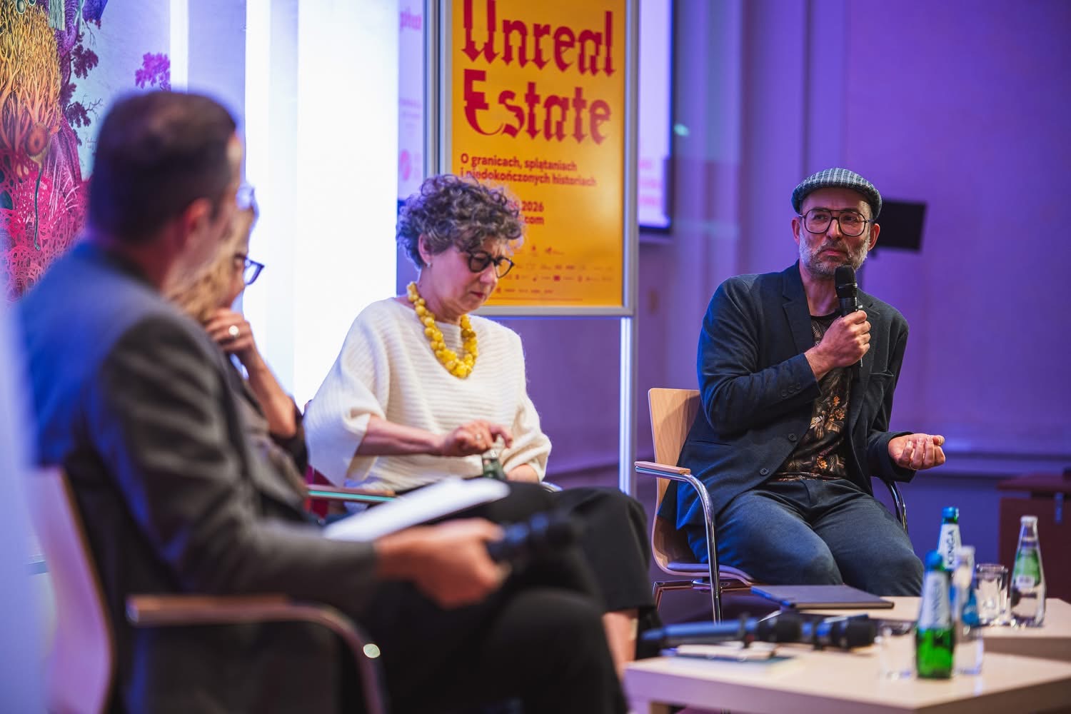 Uczestnik debaty podczas otwarcia 23. Miesiąca Fotografii w Krakowie przemawia do publiczności, trzymając mikrofon. Pozostali uczestnicy panelu siedzą obok, a w tle widoczny jest plakat projektu „Unreal Estate”.