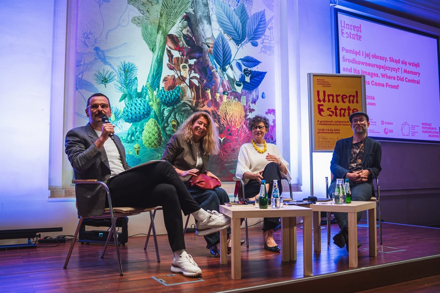 Uczestnicy debaty podczas otwarcia 23. Miesiąca Fotografii w Krakowie siedzą na scenie i rozmawiają z publicznością. W tle widoczne są plakaty projektu „Unreal Estate” oraz kolorowa dekoracja ścienna.