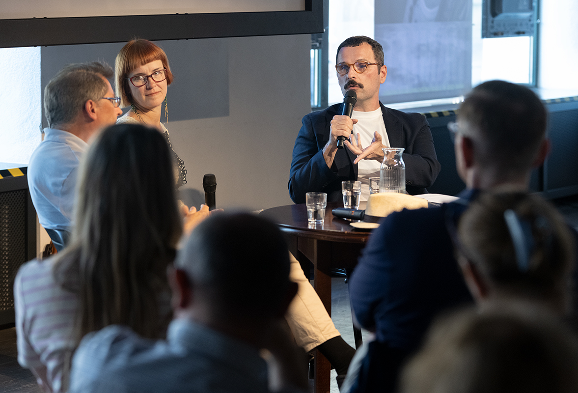 Bartosz Sadulski prowadzi dyskusję wokół 59. numeru magazynu „Herito” poświęconego Morawom. Trzymając mikrofon, zwraca się do współuczestników spotkania – Aleksandra Kaczorowskiego i dr Magdaleny Łanuszki. Rozmówcy siedzą przy niewielkim stoliku z wodą i książkami, a na pierwszym planie widoczni są słuchacze przysłuchujący się debacie. Spotkanie poświęcone jest historii, kulturze i współczesnej tożsamości Moraw.