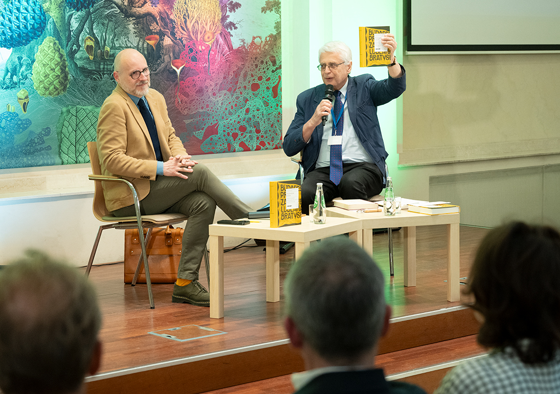 Author meeting with Dr Łukasz Galusek at the International Cultural Centre. On stage, Łukasz Galusek and Professor Jacek Purchla are seated, with Purchla holding up the book Imagined Capitals while speaking to the audience into a microphone. Tables with copies of the book and water bottles stand in front of them. Audience members are visible in the foreground.