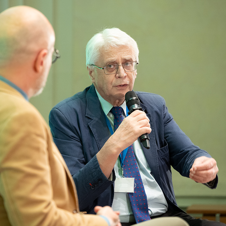 Professor Jacek Purchla speaks during an author meeting at the International Cultural Centre. He is seated, holding a microphone and gesturing while speaking. Łukasz Galusek is partially visible in the foreground.