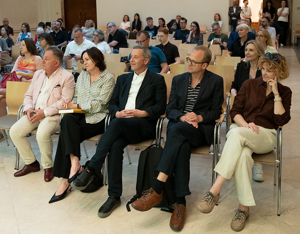 Publiczność uczestniczy w spotkaniu autorskim w sali Międzynarodowego Centrum Kultury. W pierwszym rzędzie siedzą goście wydarzenia, a pozostali uczestnicy zajmują miejsca na widowni i słuchają rozmowy odbywającej się poza kadrem.