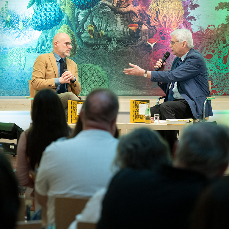 Author meeting with Dr Łukasz Galusek at the International Cultural Centre. Łukasz Galusek and Professor Jacek Purchla are seated opposite each other on stage, holding microphones and talking. Copies of the book Imagined Capitals, water bottles and books are placed on the tables in front of them. A colourful wall decoration is visible in the background.