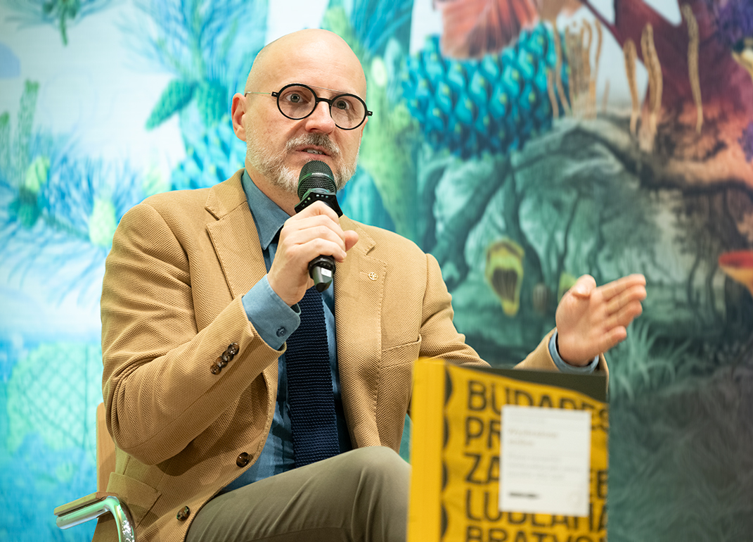 Dr Łukasz Galusek speaks during an author meeting at the International Cultural Centre. He is seated, holding a microphone and gesturing while speaking. A copy of the book Imagined Capitals is visible in the foreground. A colourful wall decoration appears in the background.