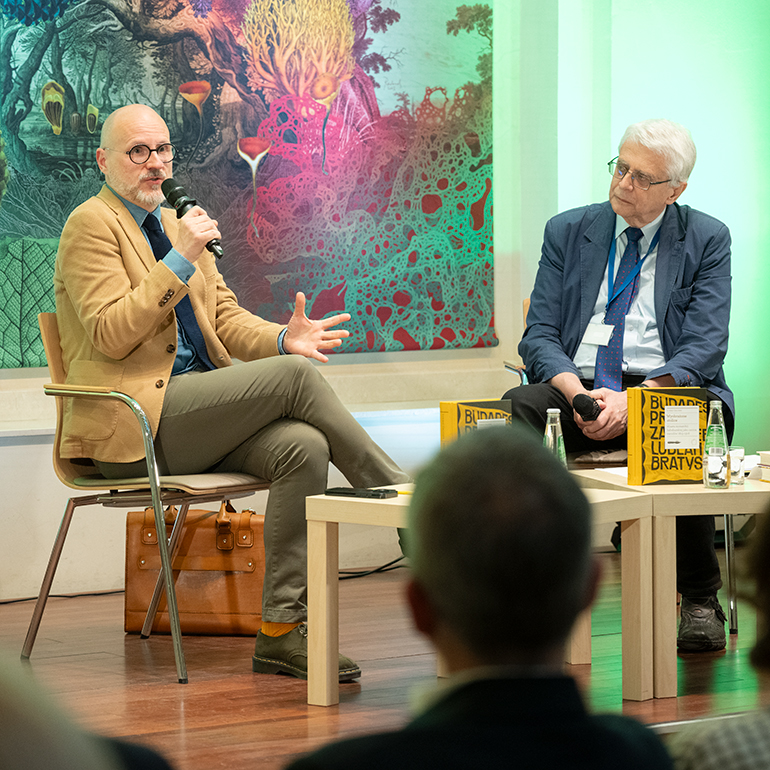 Author meeting with Dr Łukasz Galusek at the International Cultural Centre. Łukasz Galusek is seated on stage, speaking to the audience while holding a microphone and gesturing with his hand. Professor Jacek Purchla sits beside him, listening to the conversation. Copies of the book Imagined Capitals and water bottles are placed on the tables in front of them. A colourful wall decoration is visible in the background.