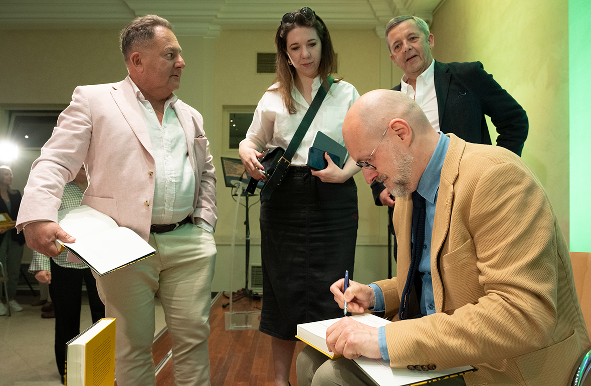 Dr Łukasz Galusek podpisuje egzemplarz książki Wyobrażone stolice po spotkaniu autorskim w Międzynarodowym Centrum Kultury. Wokół niego stoją uczestnicy wydarzenia oczekujący na autografy i rozmowę z autorem, wśród nich Robert Makłowicz.