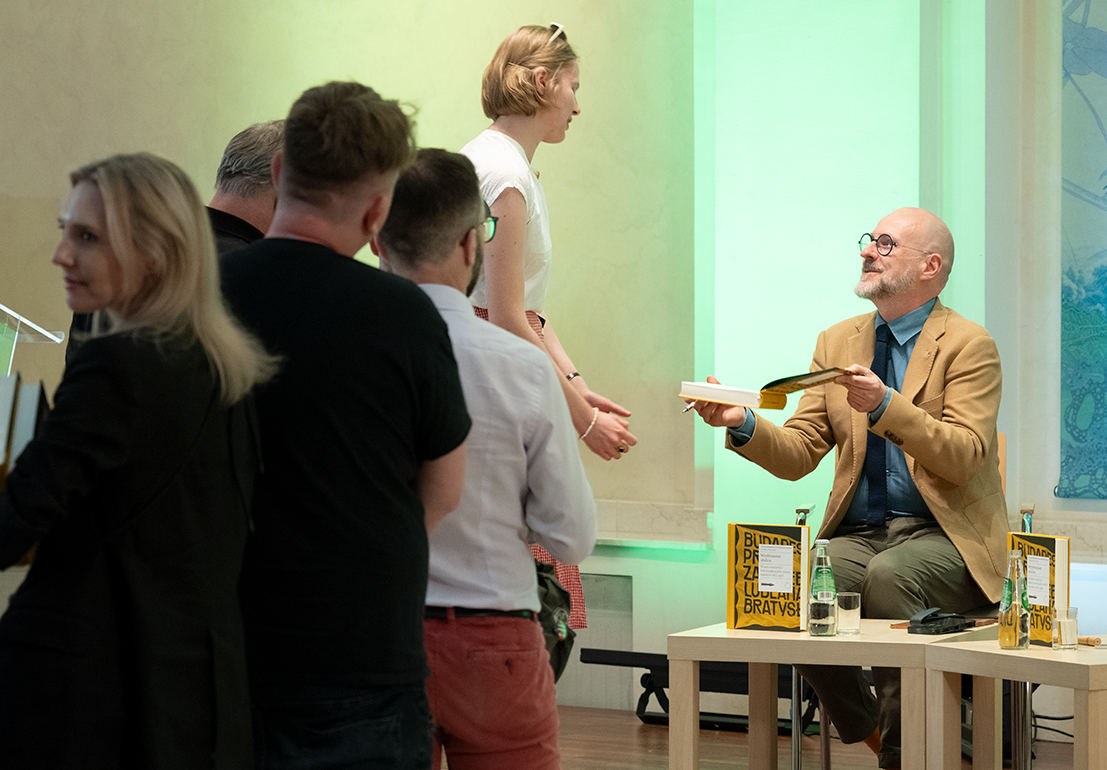 Event participants stand in line for autographs during an author meeting with Łukasz Galusek. The author sits on stage and hands a signed copy of the book “Imagined Capitals” to one of the attendees.
