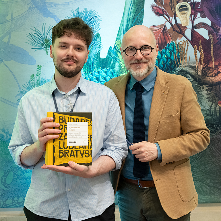 Marcin Pańtak i dr Łukasz Galusek pozują do wspólnego zdjęcia po spotkaniu autorskim w Międzynarodowym Centrum Kultury. Marcin Pańtak trzyma egzemplarz książki Wyobrażone stolice, a Łukasz Galusek stoi obok niego z długopisem w dłoni. W tle widoczna jest kolorowa dekoracja ścienna.