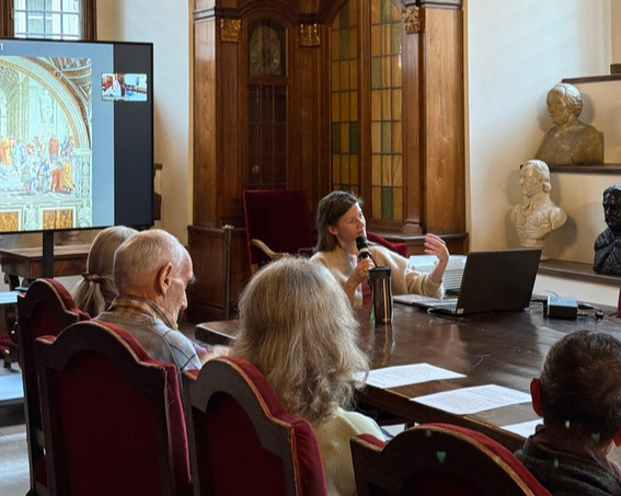 Photograph from the lecture A New View of the World: Print and Perspective. The speaker is seated at a table and addresses the audience while holding a microphone. A multimedia presentation is displayed on a screen nearby, and the interior features wooden furniture and sculptures.