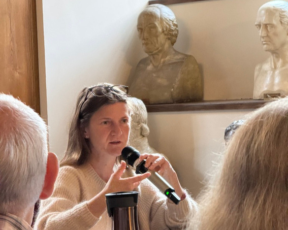 Close-up of Dr Małgorzata Jędrzejczyk during the lecture A New View of the World: Print and Perspective at Collegium Maius in Krakow. The speaker is holding a microphone and gesturing while speaking. Sculpted busts displayed in the historic interior are visible in the background.