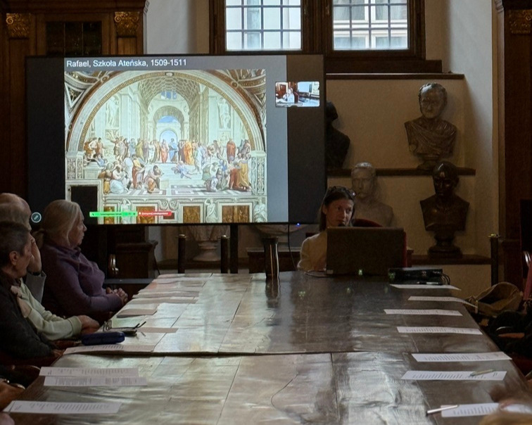 Photograph from the lecture A New View of the World: Print and Perspective at Collegium Maius in Krakow. Participants are seated around a long table while Dr Małgorzata Jędrzejczyk gives a multimedia presentation. Raphael’s painting The School of Athens is displayed on the screen.