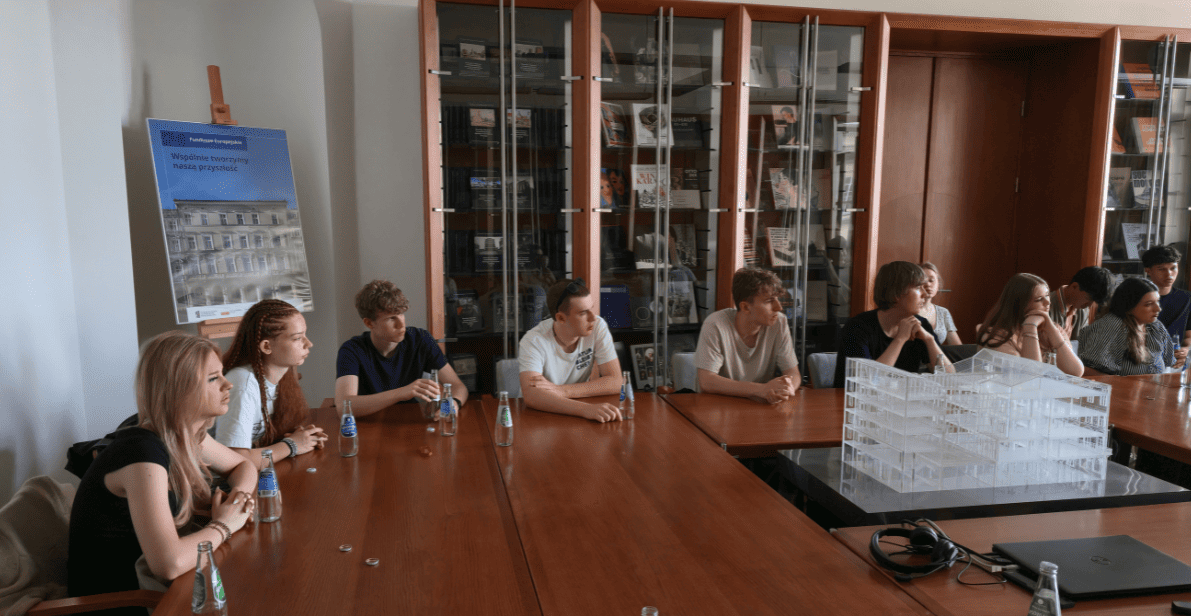 Grupa studentów uczestniczy w spotkaniu dyskusyjnym przy dużym prostokątnym stole w sali konferencyjnej lub bibliotecznej. W centrum kadru, tyłem do obiektywu, siedzi prowadząca rozmowę osoba, gestykulując podczas wypowiedzi. Wokół stołu rozmieszczonych jest kilkanaście młodych osób uważnie słuchających i biorących udział w dyskusji. Na stole znajdują się butelki wody, notatki oraz materiały robocze. W tle widoczny jest ekran multimedialny, a po bokach wysokie przeszklone regały z publikacjami. Jasne wnętrze i symetryczny układ sali podkreślają akademicki charakter spotkania.
