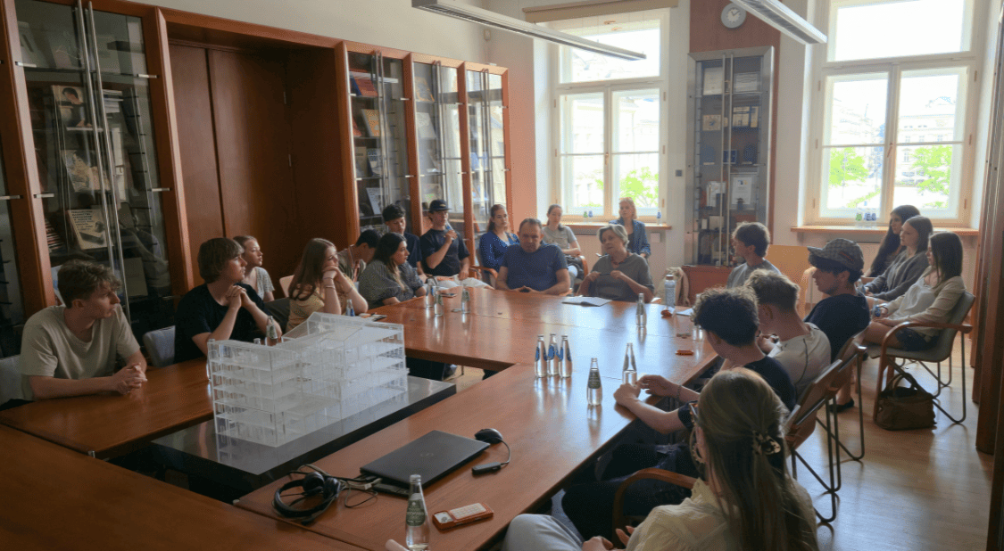 Grupa młodych osób uczestniczy w spotkaniu wokół dużego drewnianego stołu w jasnej sali bibliotecznej lub konferencyjnej. Uczestnicy siedzą po obu stronach stołu i słuchają osoby zabierającej głos w centralnej części pomieszczenia. Na stole znajdują się butelki wody, notatki, laptop, słuchawki oraz biały model architektoniczny budynku. W tle widoczne są wysokie przeszklone regały z książkami i publikacjami oraz duże okna wpuszczające naturalne światło. Spotkanie ma charakter edukacyjny i dyskusyjny.
