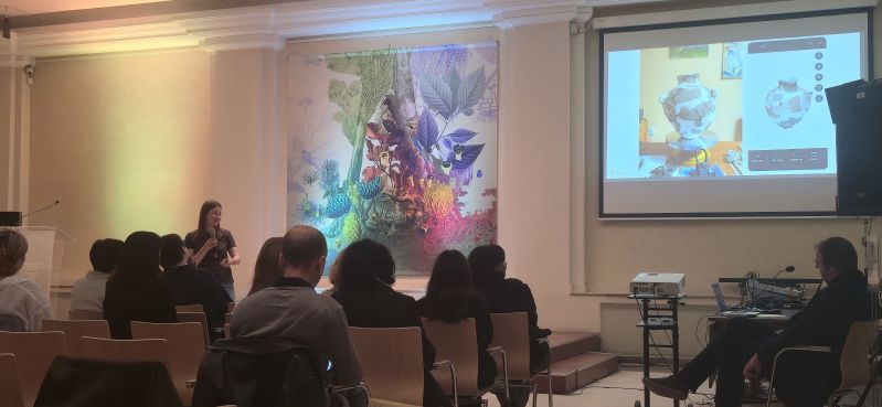 A speaker with a microphone gives a presentation to participants in the Pod Kruki Hall at the International Cultural Centre. The screen shows a photograph of a ceramic vessel alongside its digital 3D model.