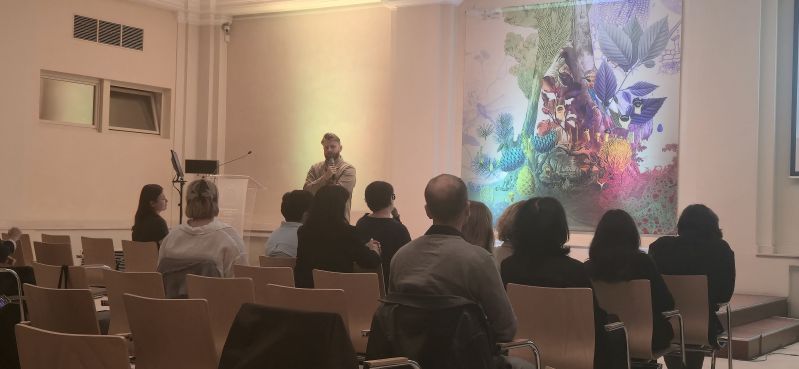 A speaker with a microphone addresses participants in the Pod Kruki Hall at the International Cultural Centre. The audience faces the stage, with a large colourful installation inspired by plant and organic forms visible in the background.