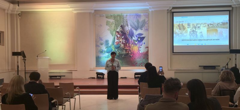 A speaker with a microphone addresses participants in the Pod Kruki Hall at the International Cultural Centre. The screen displays the website of the Berezne Local History Museum, featuring photographs related to local history and heritage.