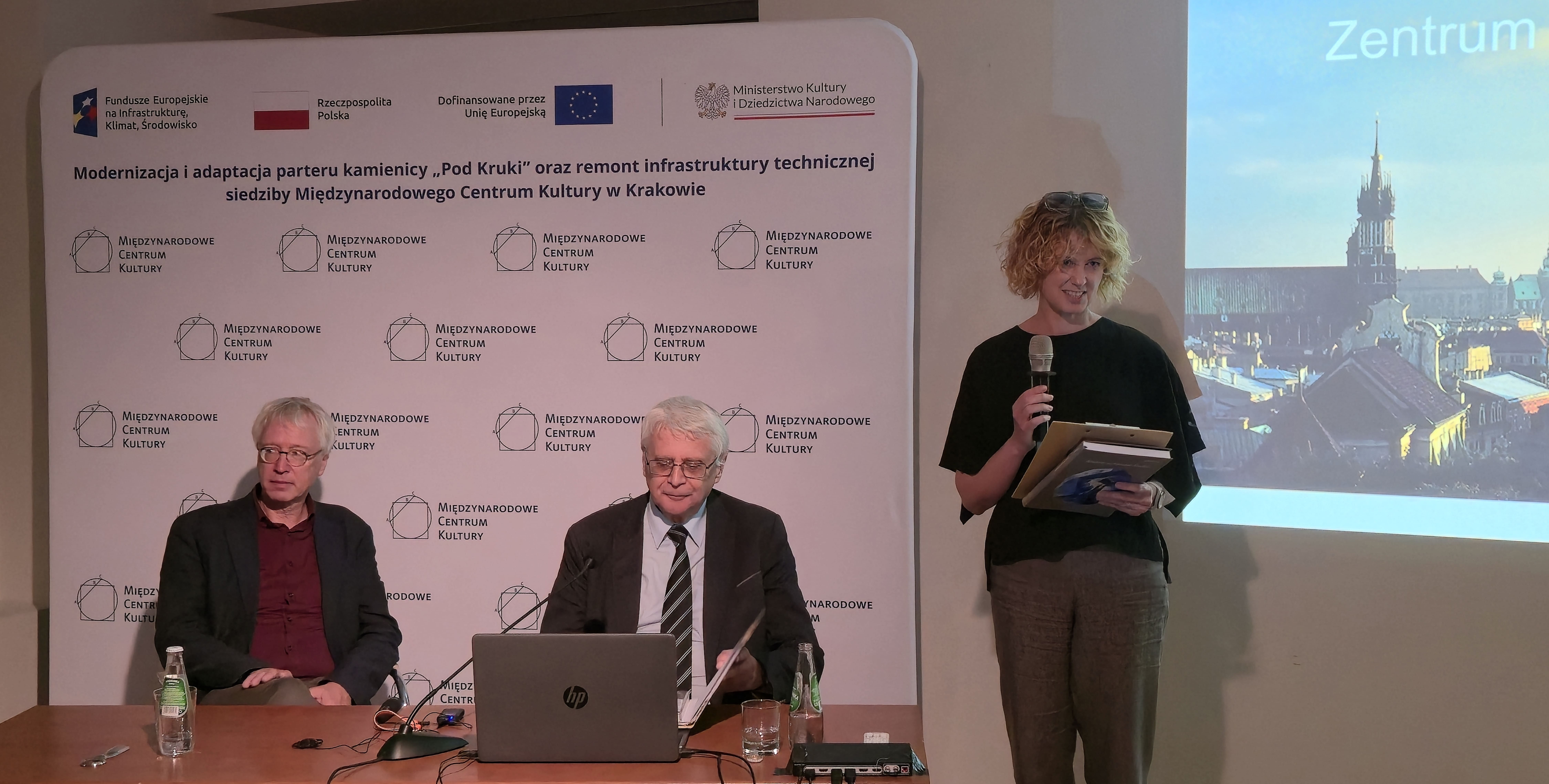 Spotkanie z delegacją z Hesji w siedzibie Międzynarodowego Centrum Kultury w Krakowie. Przed publicznością stoi Marzena Daszewska, dyrektorka programowa MCK, a przy stole siedzą prof. Jacek Purchla oraz przedstawiciel delegacji z Hesji. W tle wyświetlana jest prezentacja Krakau – Zentrum oder Peripherie? z panoramą Krakowa.