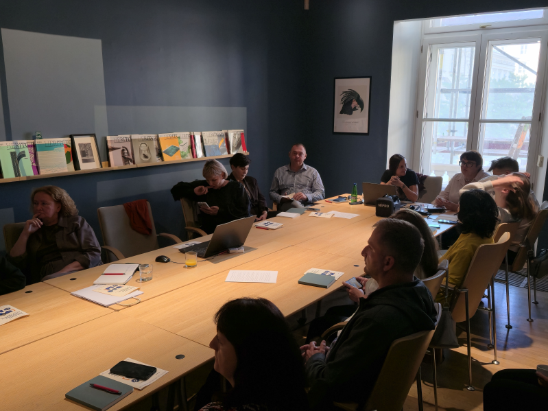 Uczestnicy i uczestniczki 4. edycji Akademii Dziedzictwa dla Ukrainy podczas spotkania przy dużym stole konferencyjnym w Międzynarodowym Centrum Kultury w Krakowie. Część osób rozmawia, inne korzystają z laptopów i telefonów lub przeglądają materiały. Na półce przy ścianie ustawione są numery kwartalnika Herito, a przez duże okno wpada światło dzienne.