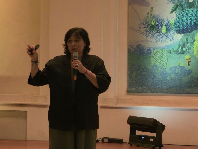 A speaker holding a microphone and presentation remote in the Pod Kruki Hall at the International Cultural Centre.