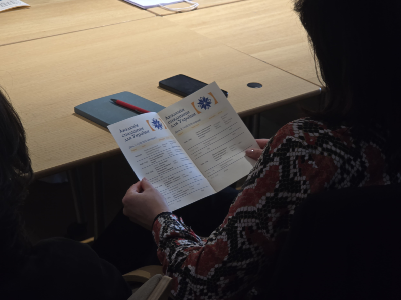 Uczestniczka 4. edycji Akademii Dziedzictwa dla Ukrainy siedzi przy stole i trzyma w rękach dwujęzyczny program Akademii. Na rozłożonej ulotce widoczny jest tytuł Akademia Dziedzictwa dla Ukrainy w języku ukraińskim oraz harmonogram zajęć. Na stole leżą notes, czerwony długopis i telefon.