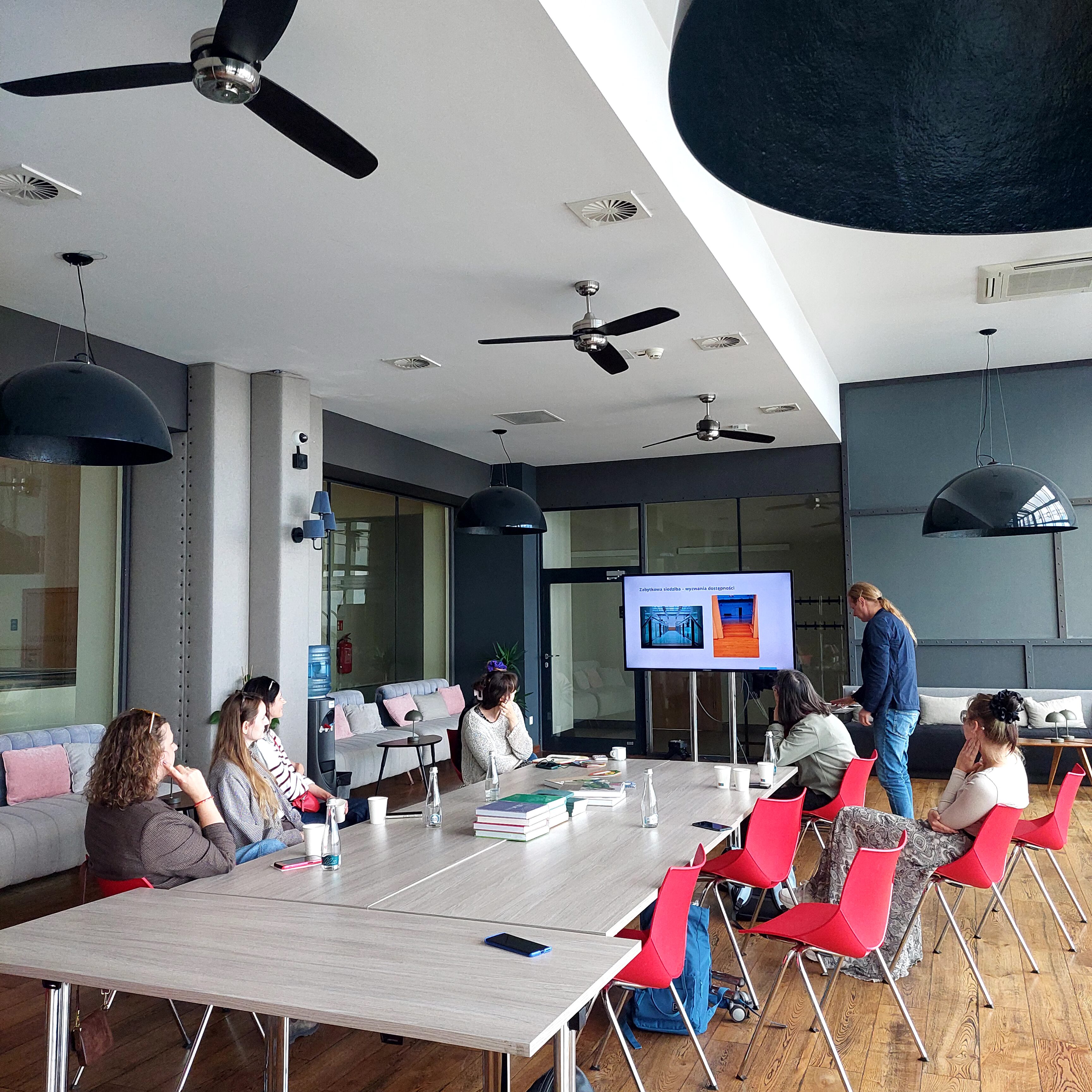 Uczestnicy wizyty studyjnej Międzynarodowego Centrum Kultury w Trójmieście podczas spotkania i prezentacji poświęconej wymianie wiedzy i doświadczeń. Grupa siedzi przy dużym stole w sali konferencyjnej, a jedna z osób prowadzi prezentację.