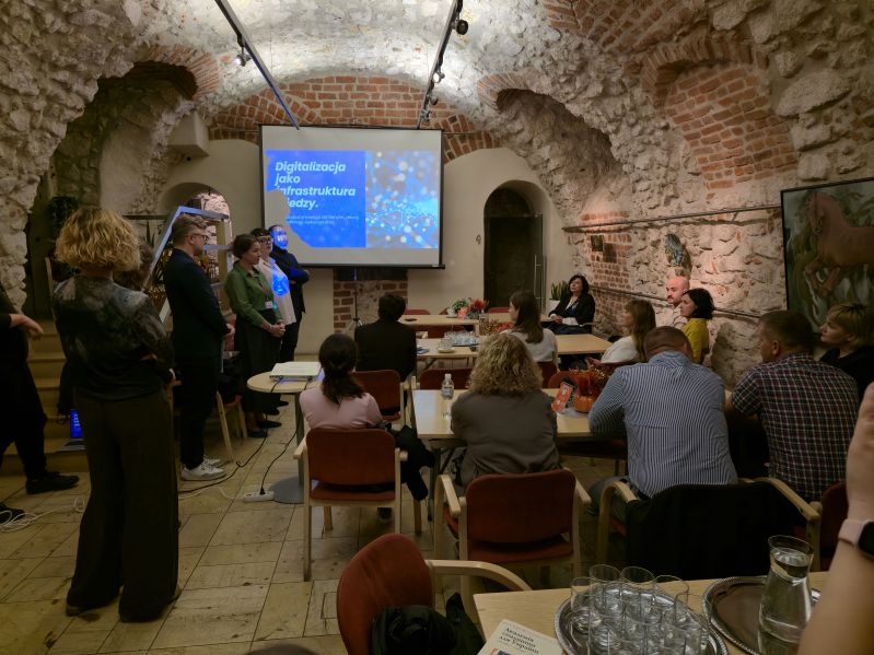 Uczestnicy IV Akademii Dziedzictwa dla Ukrainy podczas prezentacji poświęconej digitalizacji jako infrastrukturze dziedzictwa. Grupa siedzi przy stołach w zabytkowym wnętrzu z kamiennymi i ceglanymi ścianami, a prowadzący stoją obok ekranu z prezentacją.