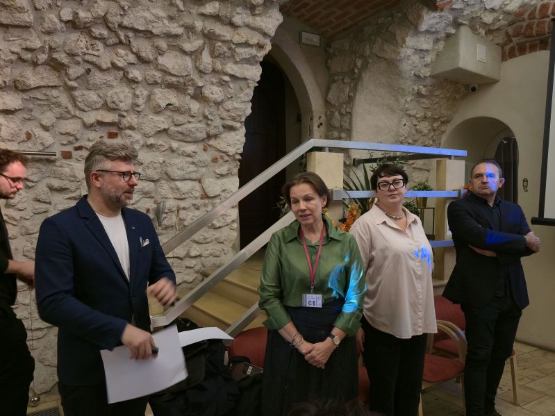 Agata Wąsowska-Pawlik wraz z uczestnikami IV Akademii Dziedzictwa dla Ukrainy podczas spotkania poświęconego sztucznej inteligencji i nowym technologiom w instytucjach kultury. Grupa stoi w zabytkowym wnętrzu z kamiennymi ścianami podczas jednej z części programu.