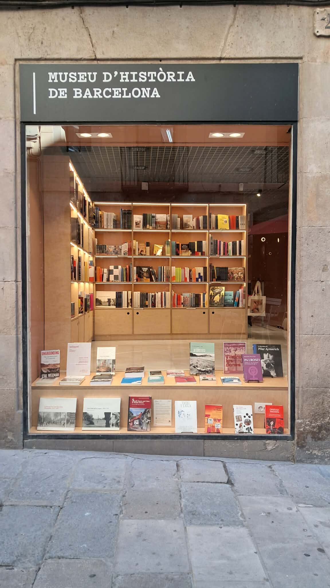 Fotografia witryny księgarni Museu d’Història de Barcelona. Za dużą przeszkloną witryną widoczne są drewniane regały wypełnione książkami oraz publikacje wyeksponowane na półkach przy oknie. Nad wejściem znajduje się szyld z nazwą muzeum.