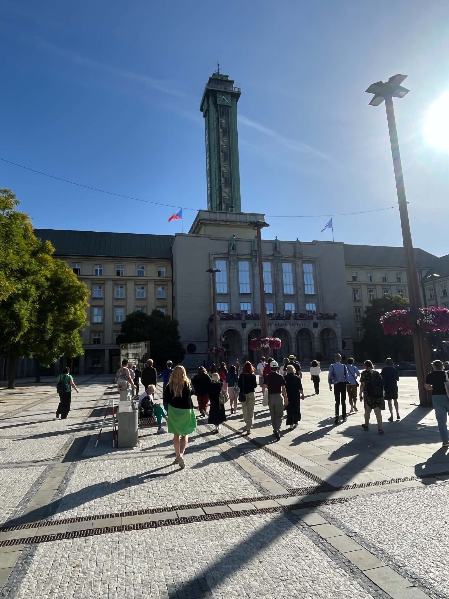 czestnicy seminarium objazdowego Europa Środkowa od środka podczas spaceru po Ostrawie w Czechach. Grupa przechodzi przez plac w kierunku monumentalnego budynku Nowego Ratusza z charakterystyczną wysoką wieżą zegarową.